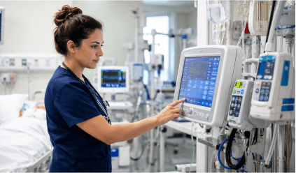 A nurse working diligently in a hospital room, with a monitor tracking patient vitals visible in the scene.
